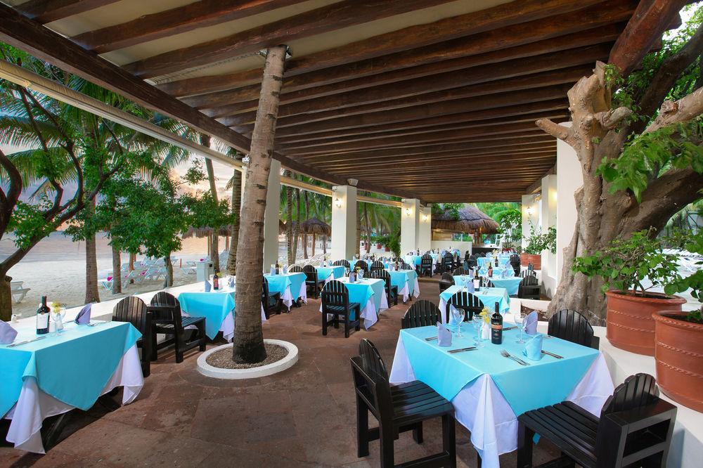 Grand Oasis Palm Hotel Cancún Kültér fotó The photo shows an outdoor dining area of a restaurant. The space features several tables covered with light blue tablecloths, each accompanied by black wooden chairs. The area is shaded by a wooden roof supported by columns, and there are leafy tree