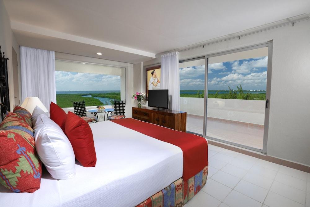 Grand Oasis Palm Hotel Cancún Kültér fotó The photo shows a modern hotel room featuring a large bed with white linens and red accents. There are decorative pillows on the bed, and a bedside table with a lamp. To the side, there’s a large window with white curtains that offers a view of a bal