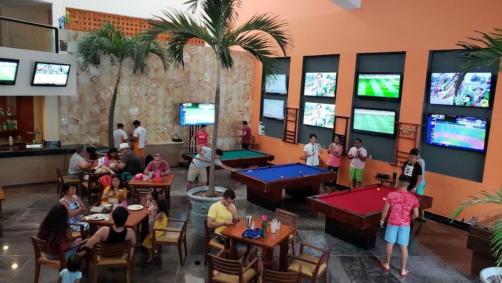 Grand Oasis Palm Hotel Cancún Kültér fotó The photo shows a lively recreational area inside a building, likely a bar or game room. There are several pool tables, one blue and one red, where people are playing or waiting for their turn. 

Around the area, groups of people are seated at tables