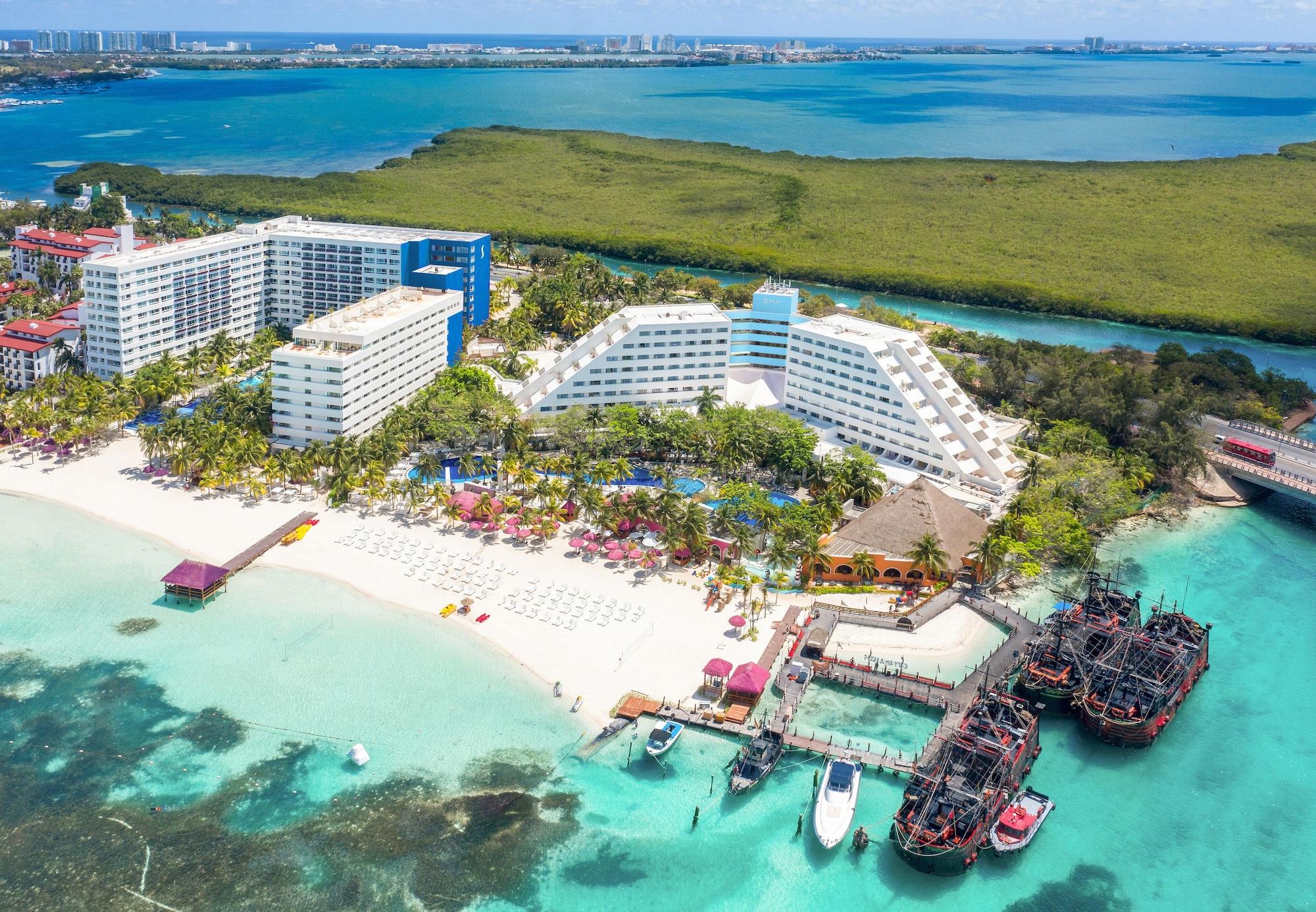 Grand Oasis Palm Hotel Cancún Kültér fotó The photo shows a beautiful coastal resort area. In the foreground, there is a sandy beach with lounge chairs and umbrellas, while the clear turquoise waters are visible. The resort features large white and blue buildings, with lush greenery surround
