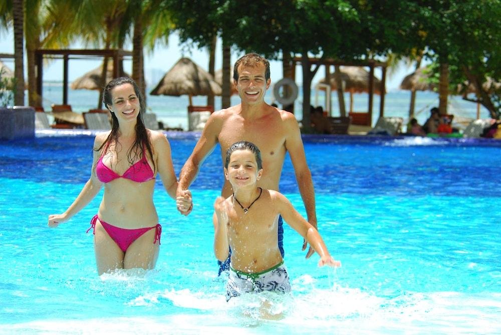 Grand Oasis Palm Hotel Cancún Kültér fotó The photo depicts a family enjoying a day at a pool. A woman in a pink bikini and a man are holding hands, while a boy runs happily in the water beside them. The pool is bright blue, and the background features palm trees and thatched huts, suggestin