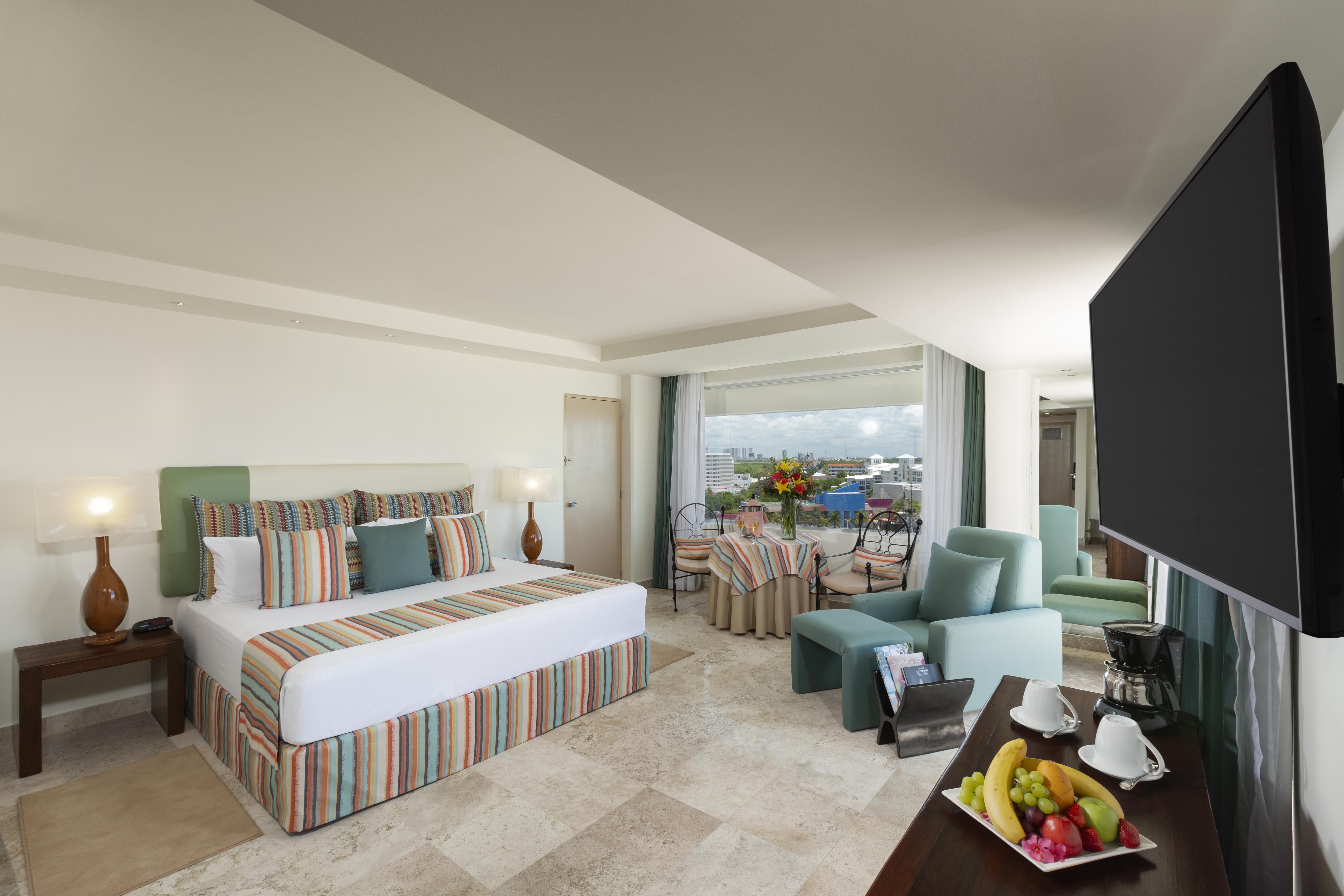 Grand Oasis Palm Hotel Cancún Kültér fotó The photo shows a spacious hotel room with modern decor. It features a large bed with a striped duvet and decorative pillows, flanked by two lamps. There are comfortable seating areas with light green chairs and a small table. A balcony can be seen i