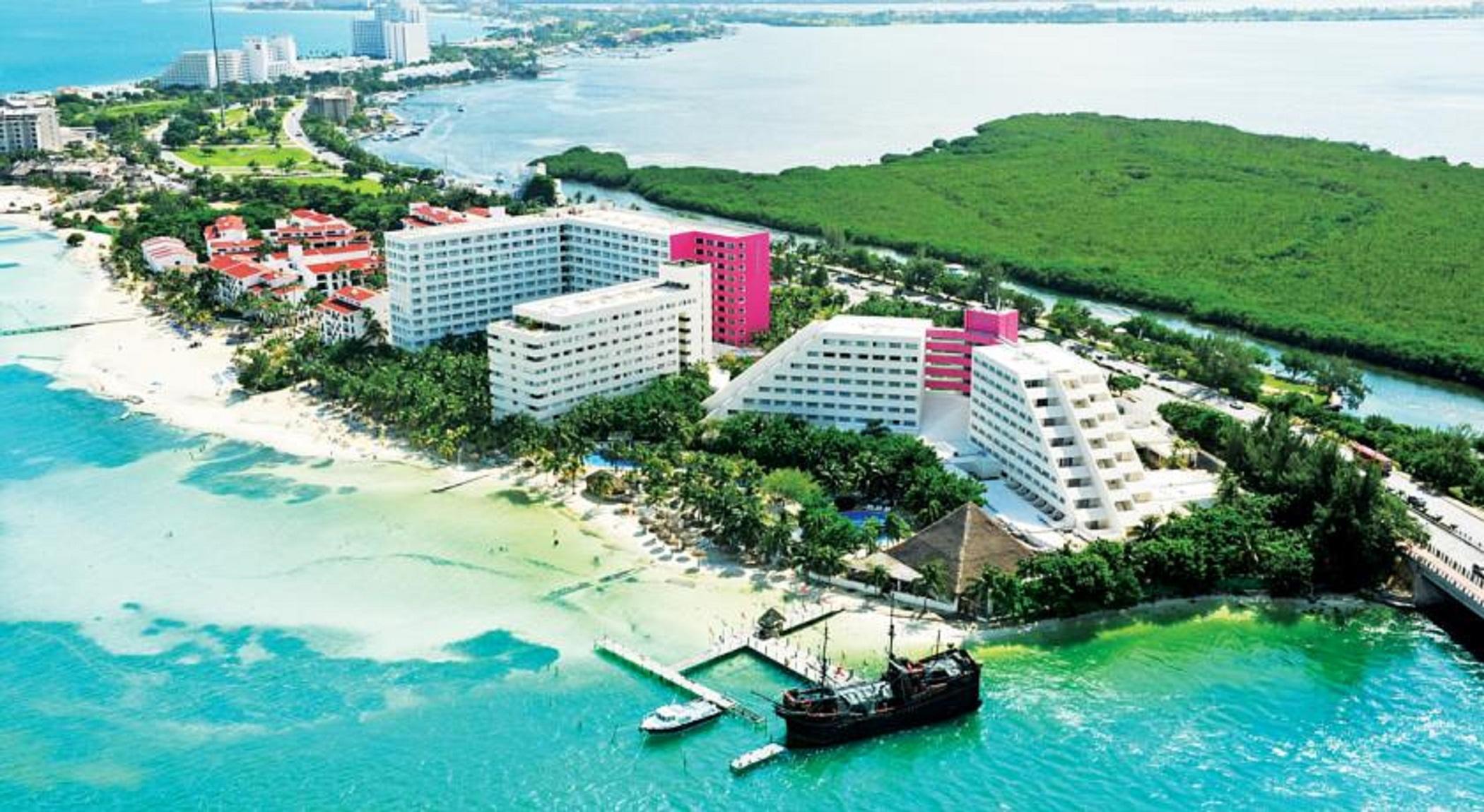 Grand Oasis Palm Hotel Cancún Kültér fotó The photo shows a coastal resort area featuring several large hotel buildings, some of which are white and others are pink. The hotels are situated near a sandy beach, with clear blue water stretching out into the background. There are palm trees and