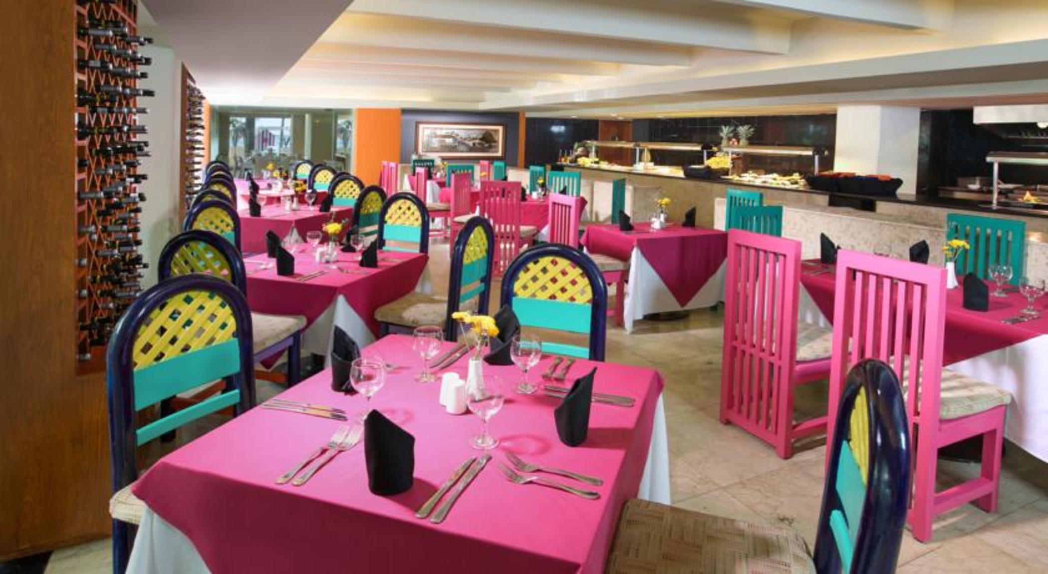 Grand Oasis Palm Hotel Cancún Kültér fotó The photo shows a vibrant restaurant interior featuring colorful decor. There are multiple tables covered with bright pink tablecloths. Each table is equipped with utensils, napkins, and a small centerpiece, possibly a flower. The chairs are painted 