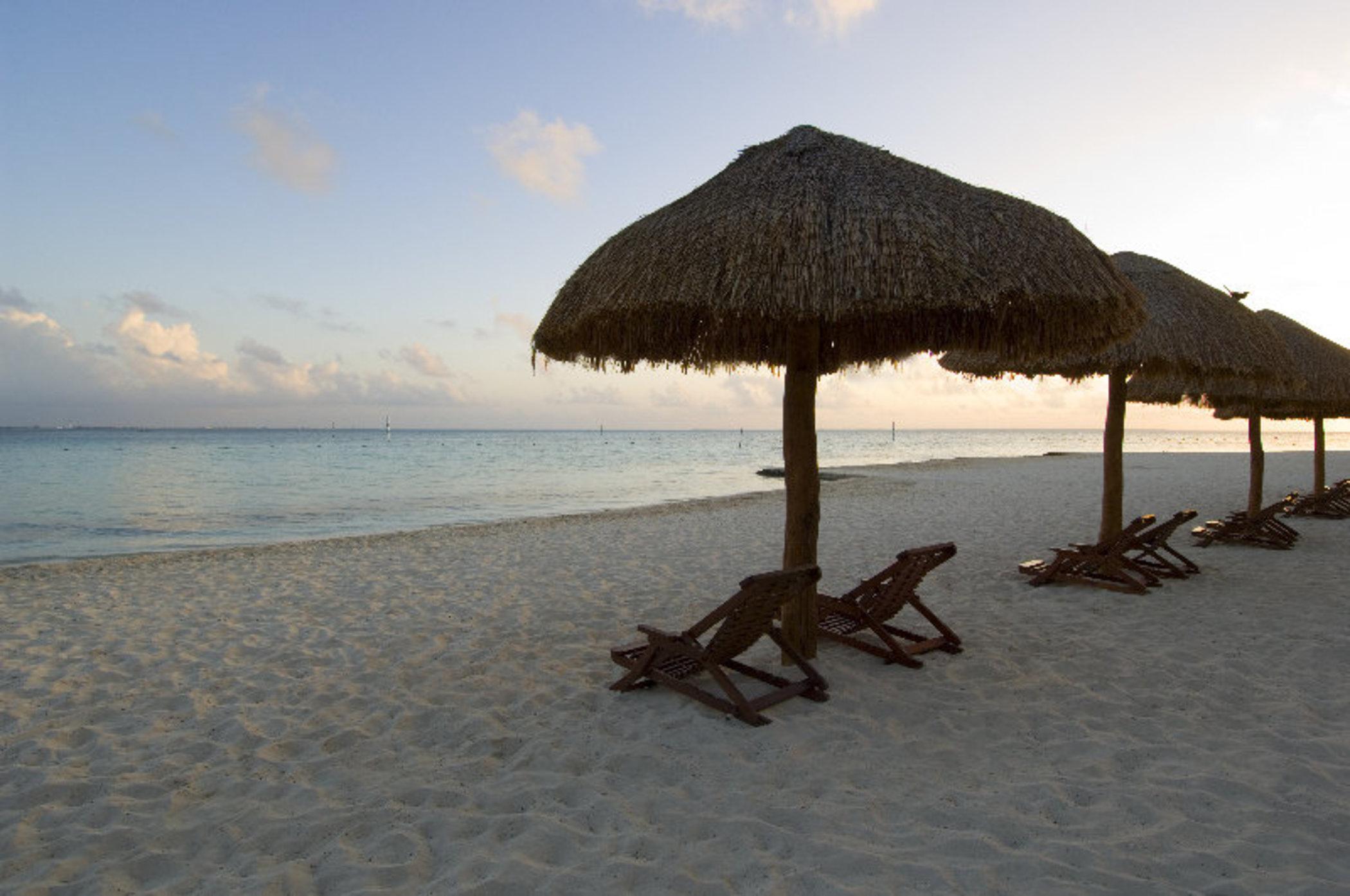 Grand Oasis Palm Hotel Cancún Kültér fotó The photo depicts a serene beach scene featuring several thatched umbrellas on the sand. Beneath the umbrellas are wooden lounge chairs, positioned for relaxation. The beach has soft, white sand, and the calm waters of the sea reflect the colors of t