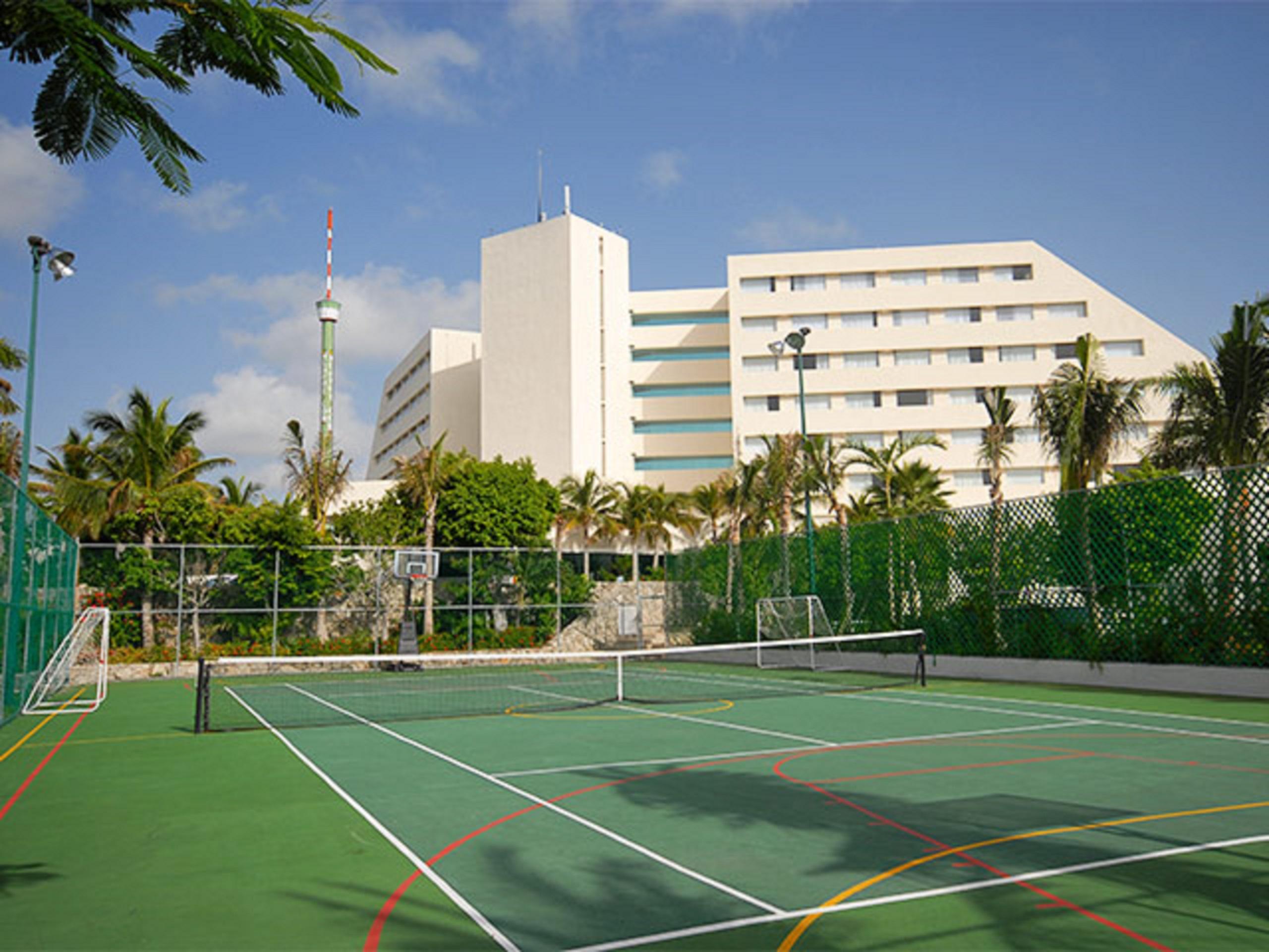 Grand Oasis Palm Hotel Cancún Kültér fotó The photo shows a sports facility featuring a tennis court in the foreground. The court is surrounded by a green fence and has distinctive lines marking the playing area. In the background, there is a tall, modern building, possibly a hotel or resort