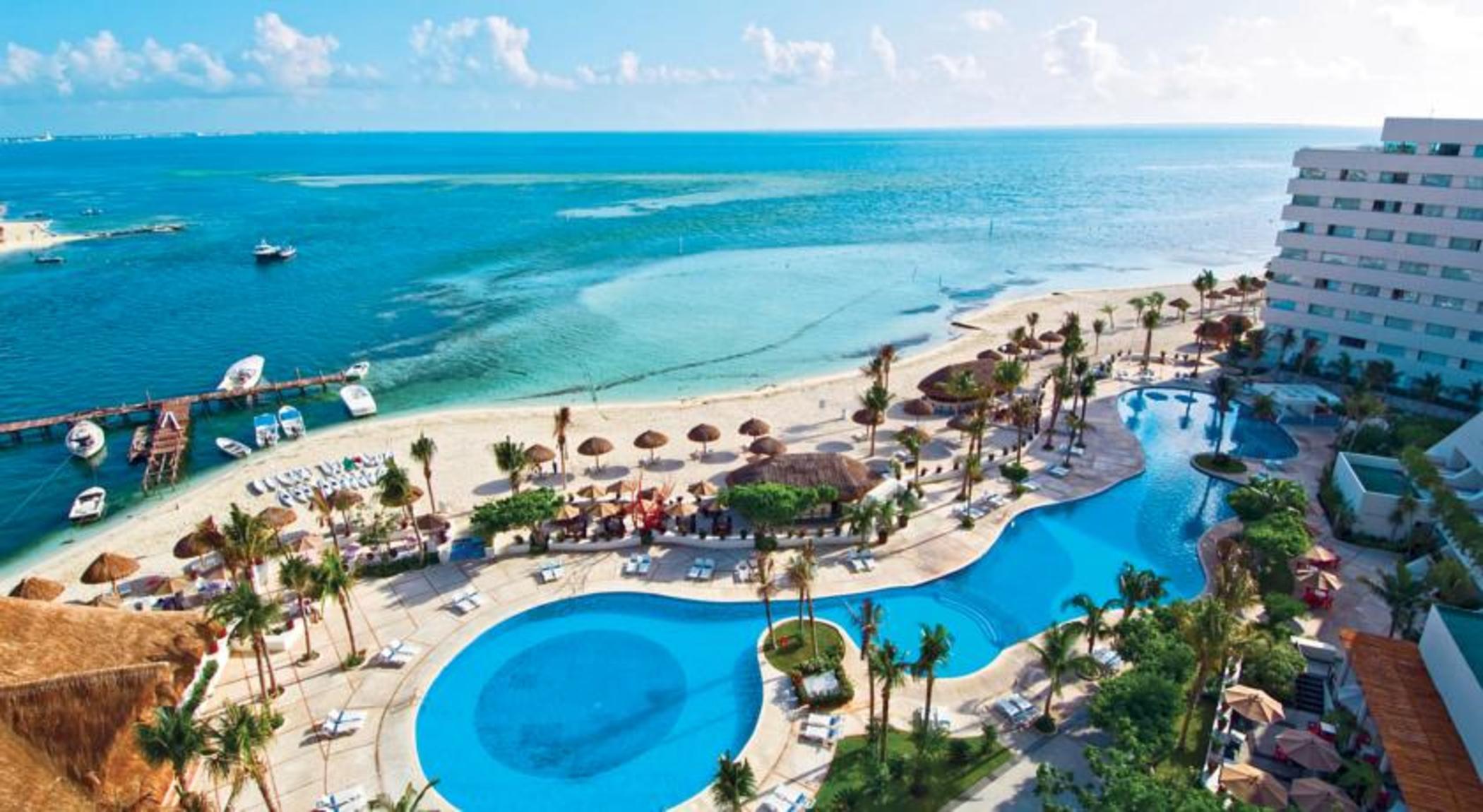 Grand Oasis Palm Hotel Cancún Kültér fotó The photo depicts a beautiful resort area located near a beach. In the foreground, there are two large swimming pools, one of which features a curved shape, surrounded by lounge chairs. Palm trees and tropical plants are scattered throughout the area