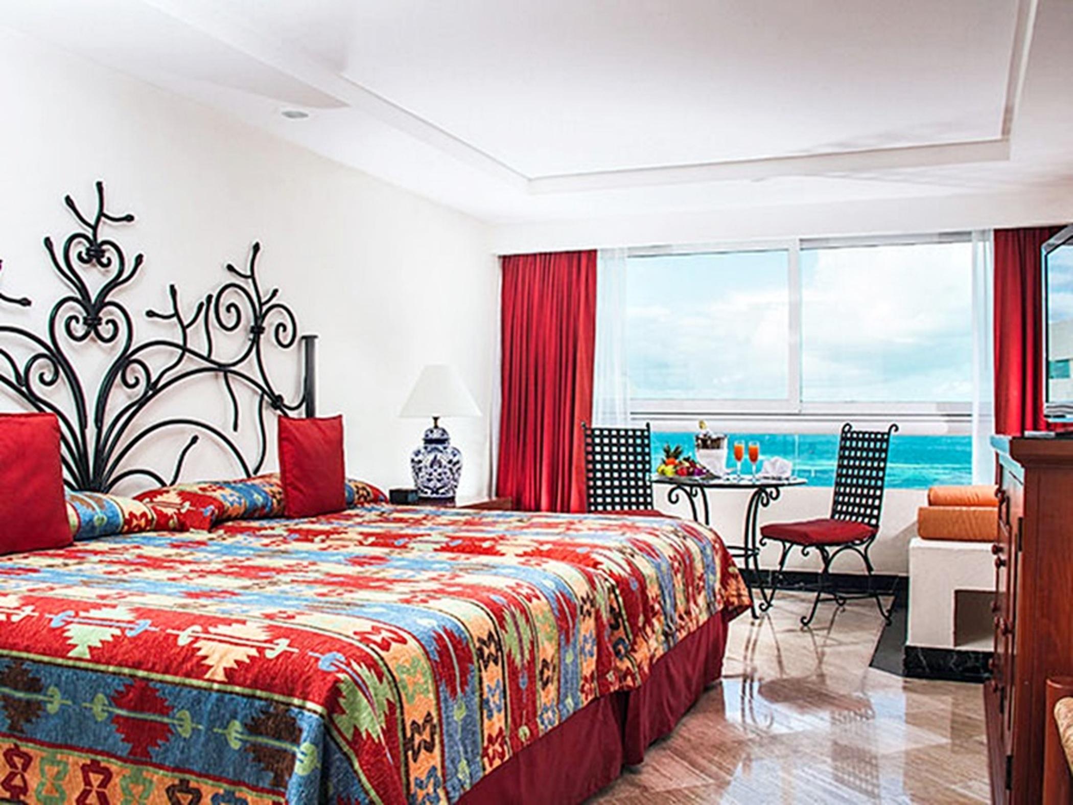 Grand Oasis Palm Hotel Cancún Kültér fotó The photo shows a well-decorated hotel room featuring two large beds with vibrant, patterned bedding. The headboards are stylishly designed, and the room has colorful accents like red curtains and cushions. A small table with chairs is positioned by 