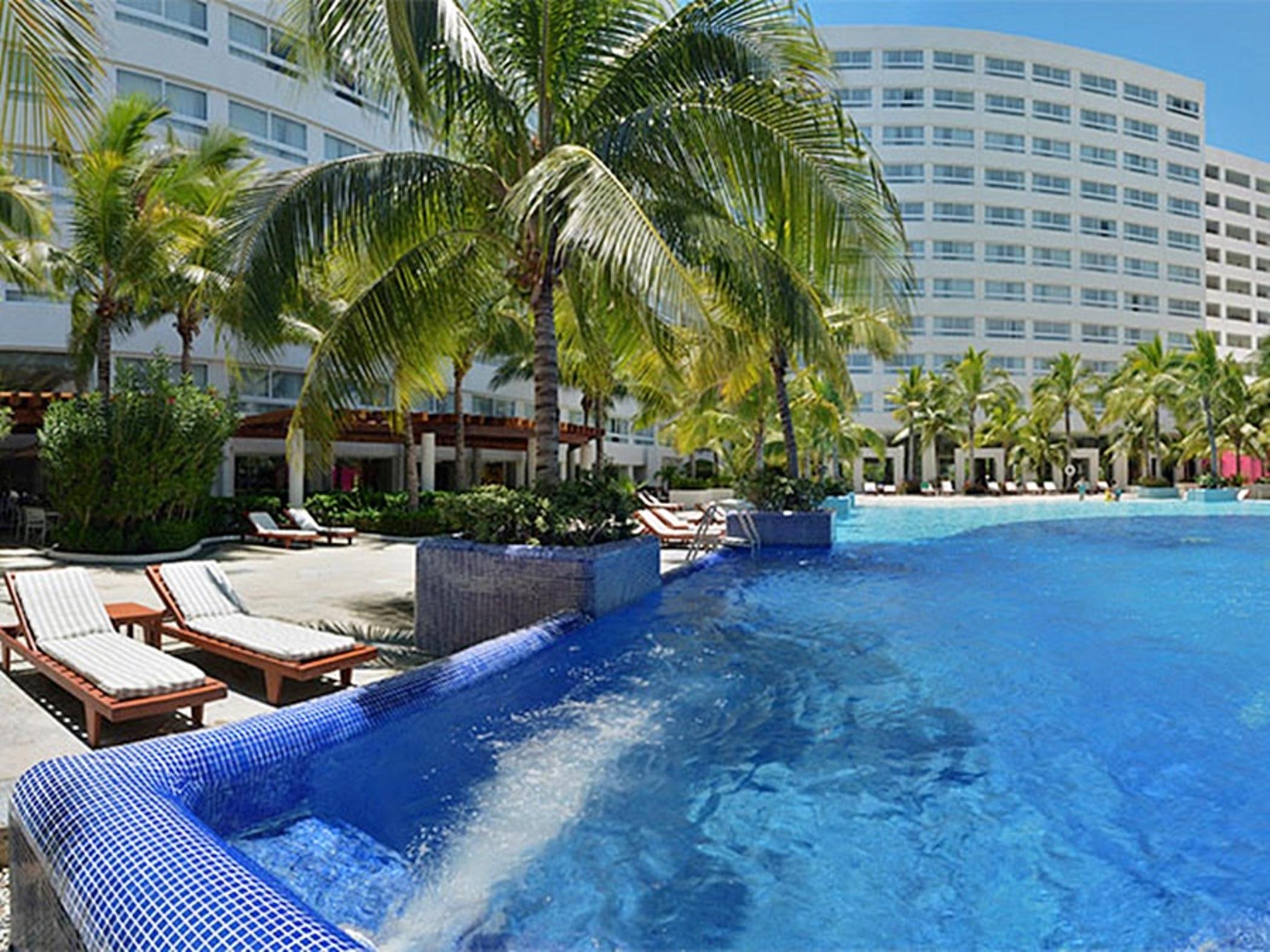 Grand Oasis Palm Hotel Cancún Kültér fotó The photo shows a resort-style setting featuring a large swimming pool with clear blue water. Surrounding the pool are lounge chairs, some of which are positioned under palm trees, providing a tropical ambiance. In the background, there is a multi-st