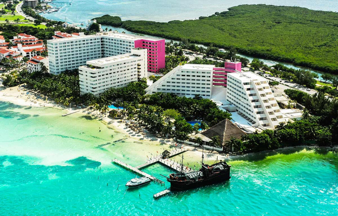 Grand Oasis Palm Hotel Cancún Kültér fotó The photo shows an aerial view of a resort area located near a beach. There are several large buildings, with a mix of white and pink architecture. The buildings are surrounded by lush greenery and palm trees, creating a tropical atmosphere. 

In the